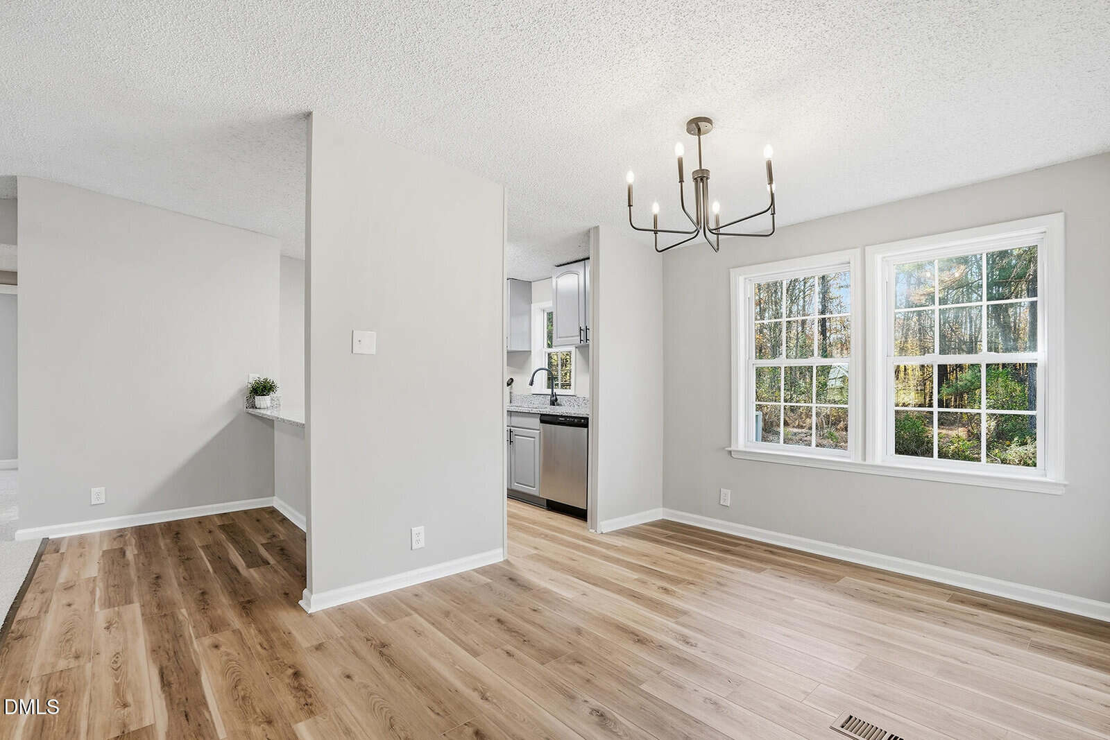 A room with light wood flooring, light gray walls, a modern black chandelier, and a large window. The space opens into a kitchen area with white cabinets and stainless steel appliances.