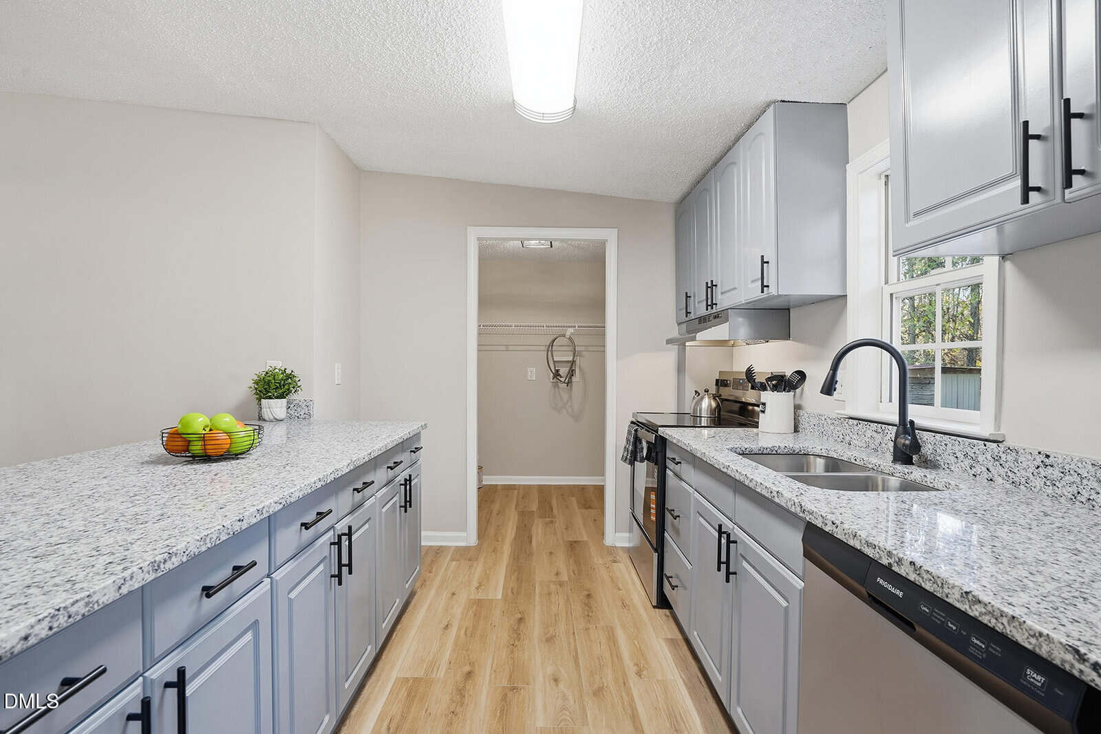 Modern kitchen with light gray cabinets, granite countertops, stainless steel appliances, wood-look flooring, under-cabinet lighting, and a bowl of fruit on the counter. A pantry is visible through an open doorway.