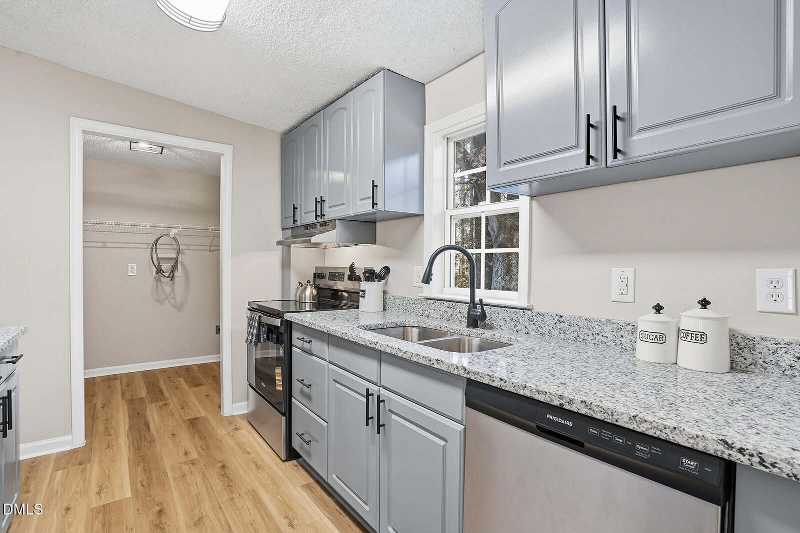 Modern kitchen with light gray cabinets, granite countertops, stainless steel appliances, wood-style flooring, and a window above the sink. A laundry area with shelving is visible through an open door.