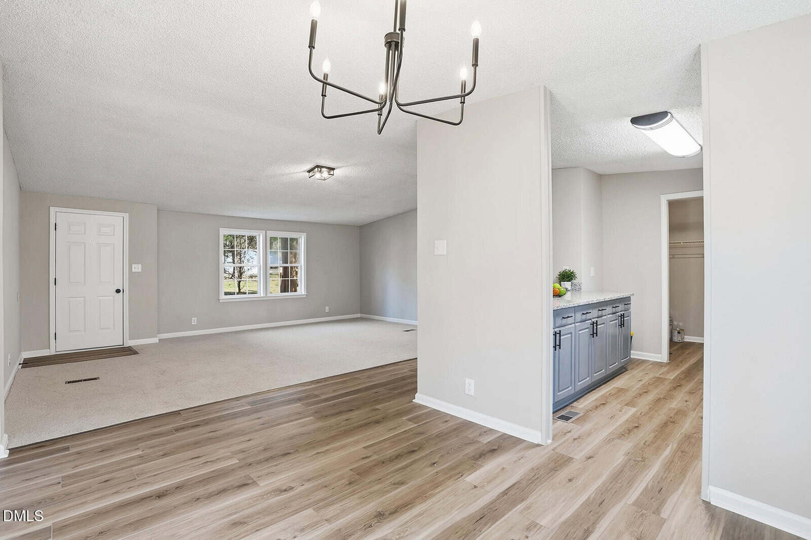 Open living area with light wood flooring, neutral walls, a modern chandelier, carpeted section, front door, large window, and a view of a hallway with blue cabinets and countertop.