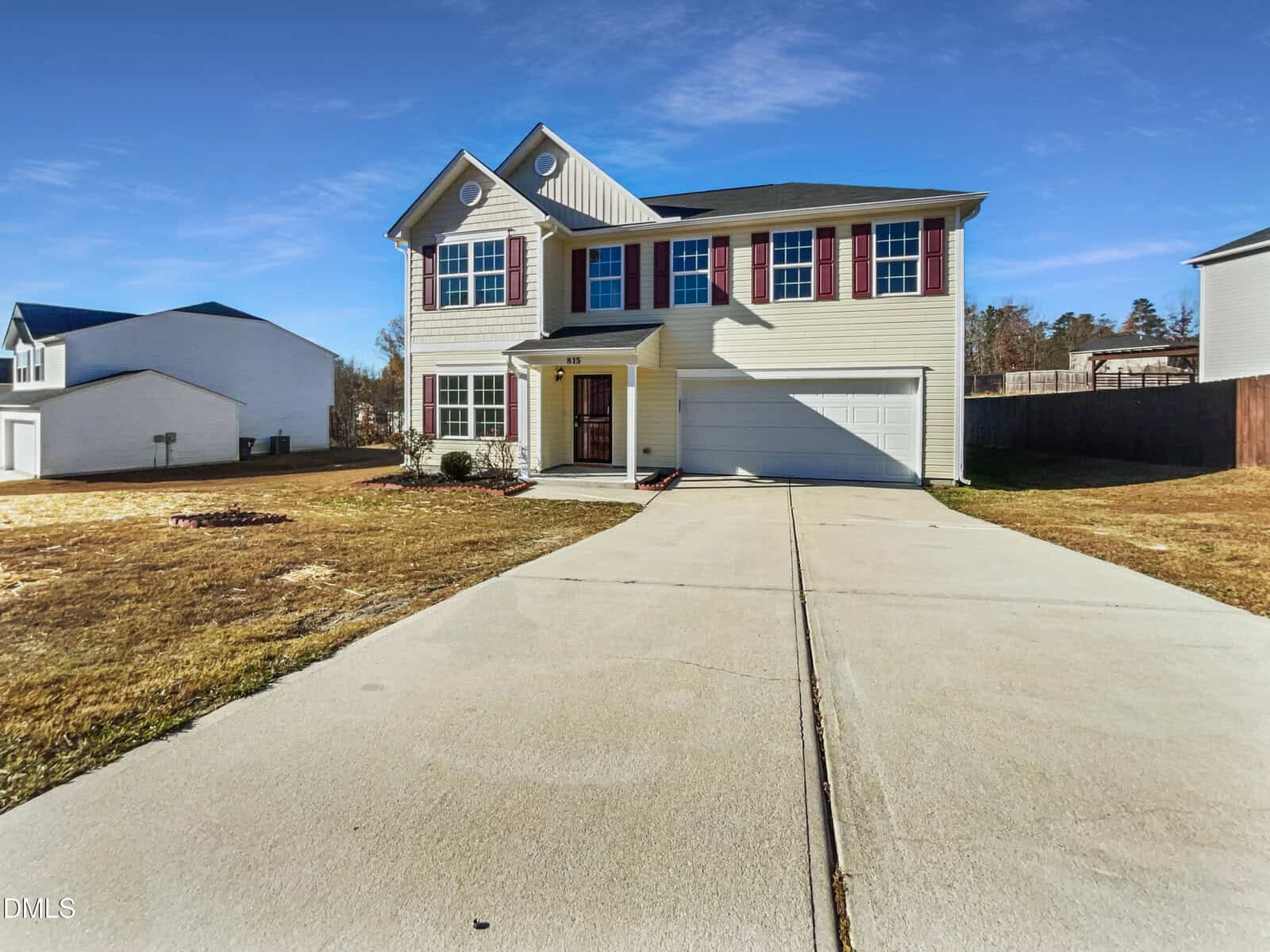 Two-story beige house with red shutters, white trim, and a two-car garage. The house has a long concrete driveway, a small front porch, and a large front yard with minimal landscaping. Clear blue sky in the background.