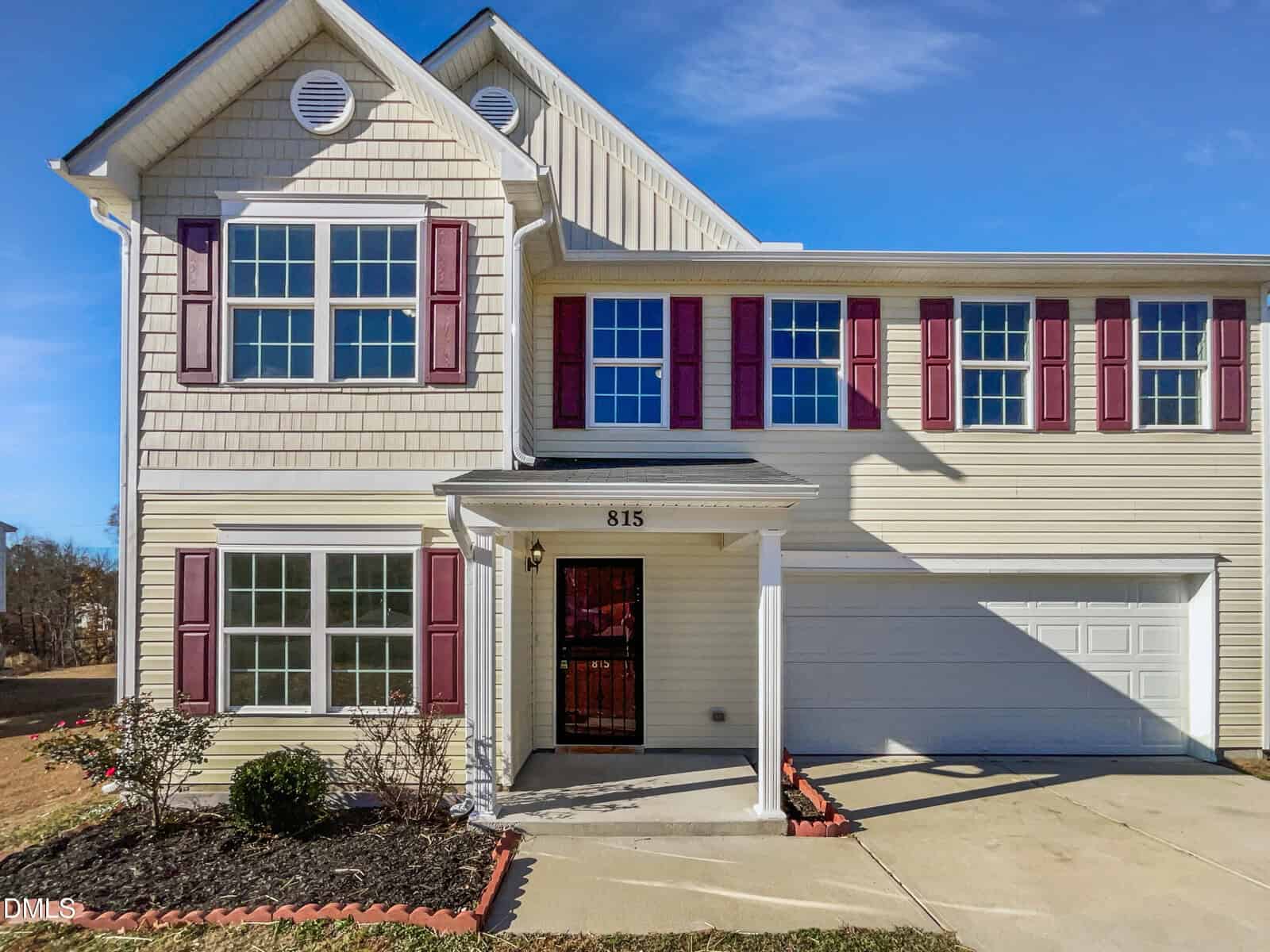 Charming two-story beige house at 815 Pitch Pine Drive, Creedmoor, with red shutters, white trim, a covered front entrance, and an attached two-car garage. Landscaped bushes border the walkway; driveway and lawn are visible.