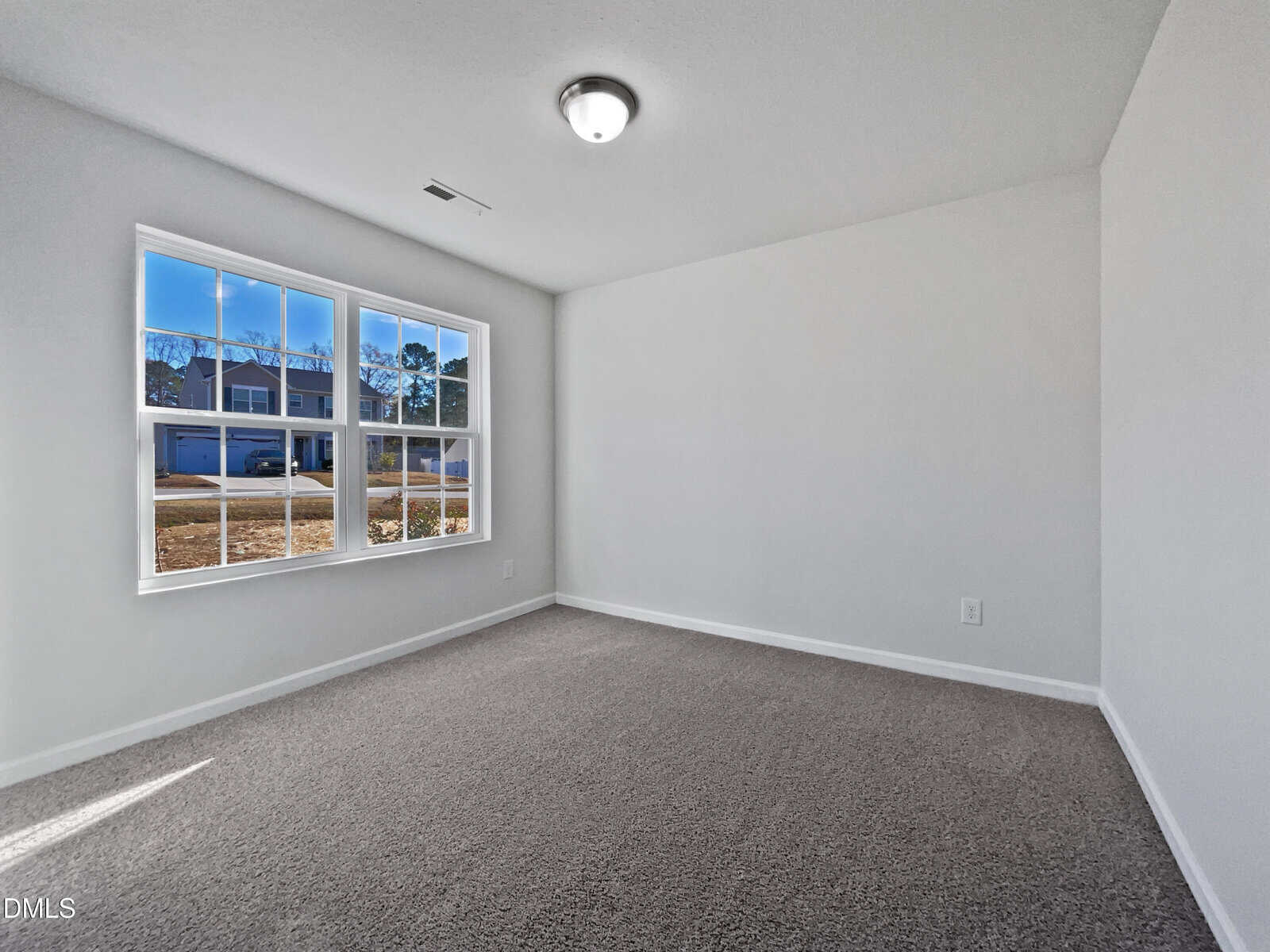 Bright, empty room with white walls, gray carpet, large window letting in natural light, and a ceiling light fixture. Houses and trees are visible outside the window.
