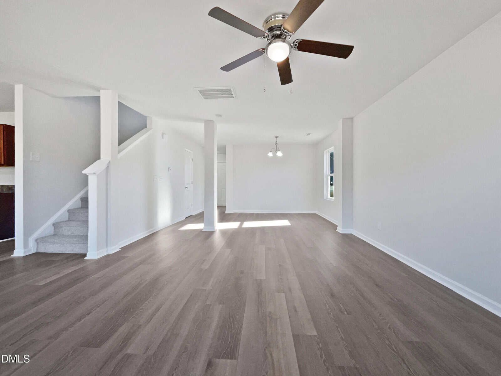 Spacious living area with light gray walls, wood-look flooring, ceiling fan with lights, and white staircase. Two pendant lights hang near a window, and natural light enters from multiple angles.