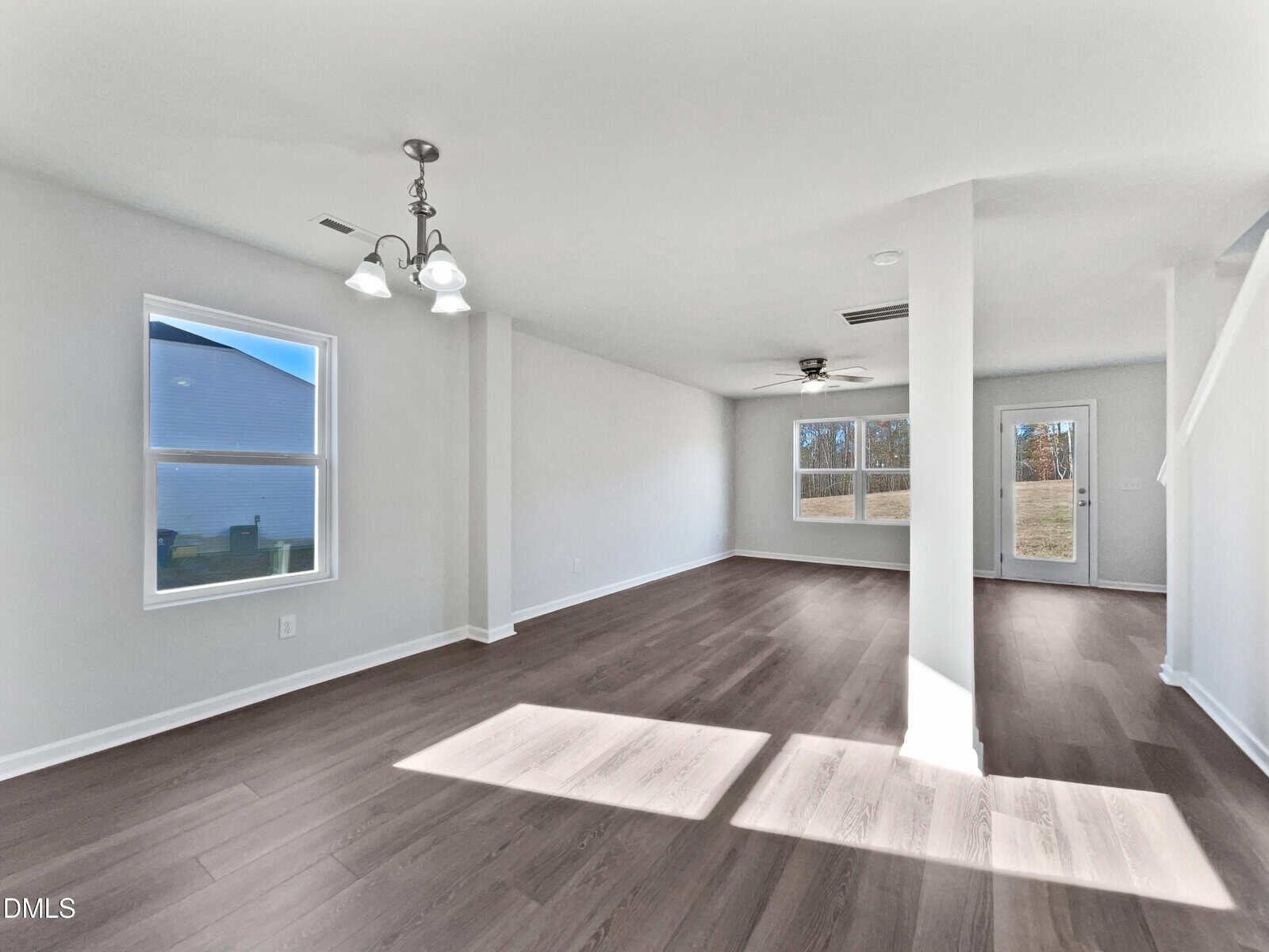 A spacious, empty room with light-colored walls, wood flooring, multiple windows letting in natural light, a ceiling fan, and a staircase to the right. The room has a clean, modern appearance.