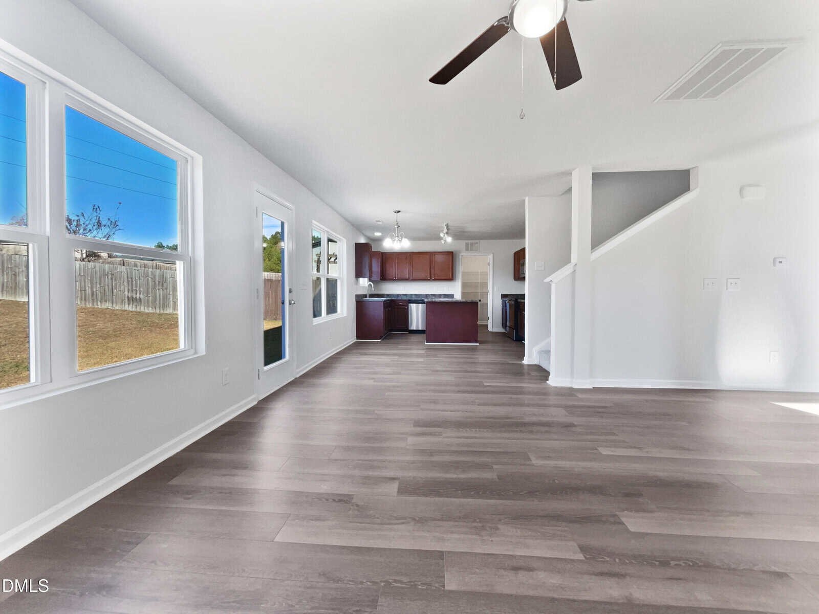 A spacious, empty open-plan living and dining area with light wood floors, large windows, a ceiling fan, and a view of the kitchen in the background; stairs lead to the upper floor on the right.