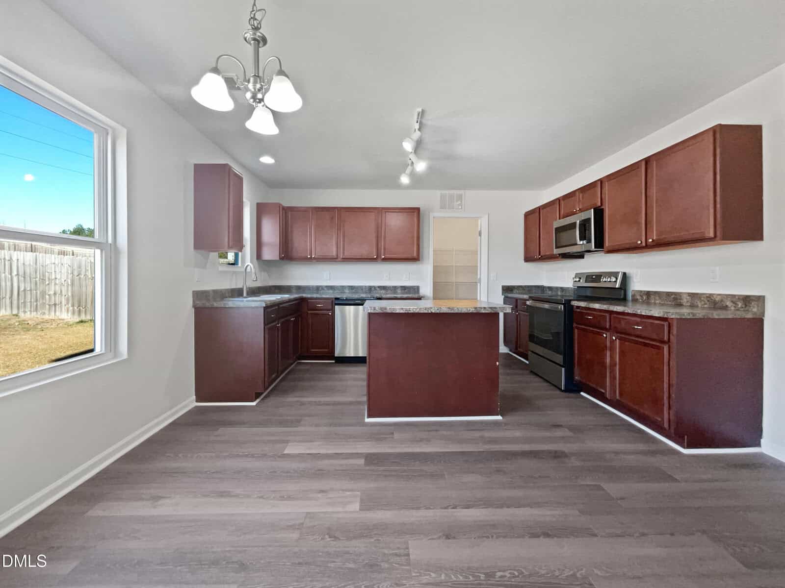 A modern kitchen with dark wood cabinets, stainless steel appliances, an island in the center, overhead lighting, and large window providing natural light. The floor is a light wood laminate.