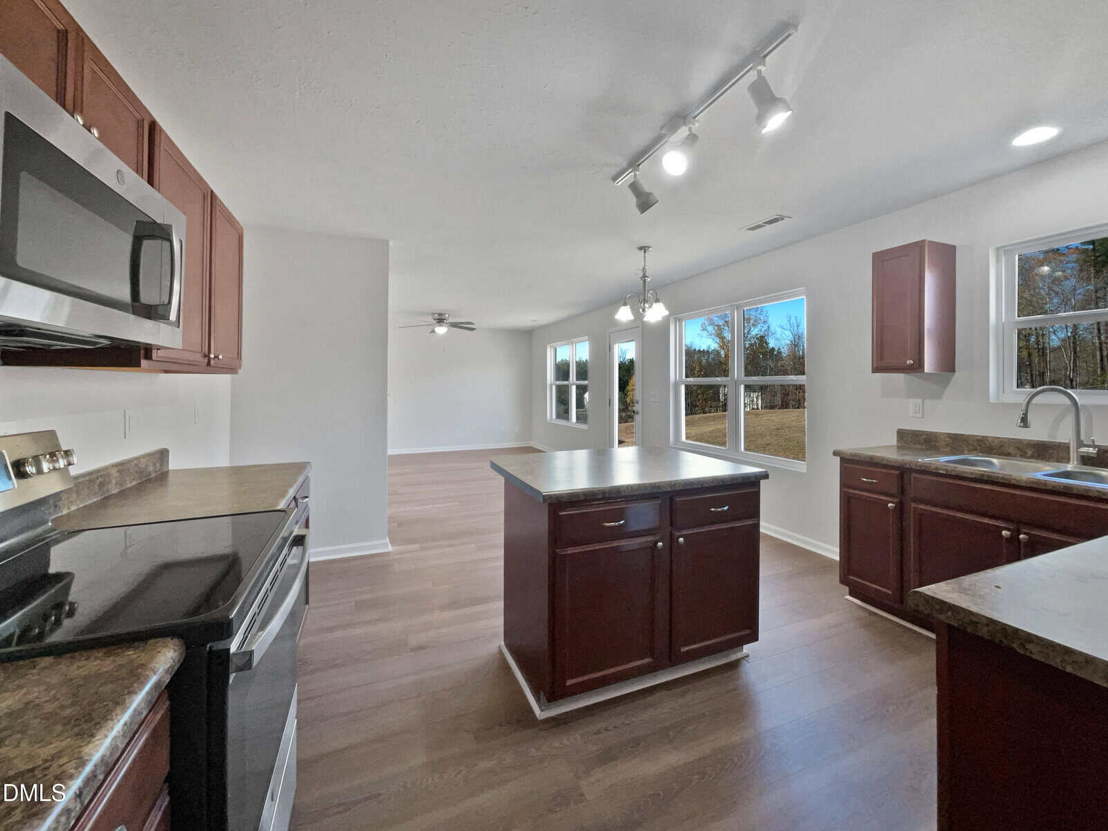 A modern kitchen with dark wood cabinets, stainless steel appliances, a central island, and adjoining dining area. Large windows provide natural light and offer views of an open grassy yard.