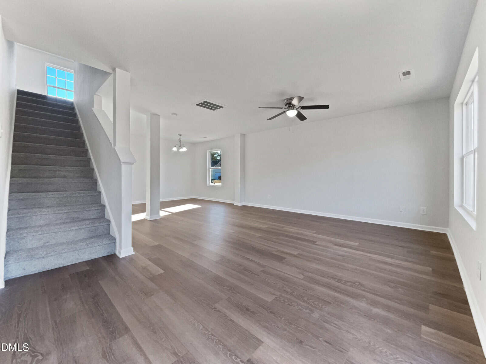 A spacious, empty living room with light-colored walls, wood flooring, a ceiling fan, multiple windows, and a carpeted staircase on the left side. Natural light enters the room from the windows.