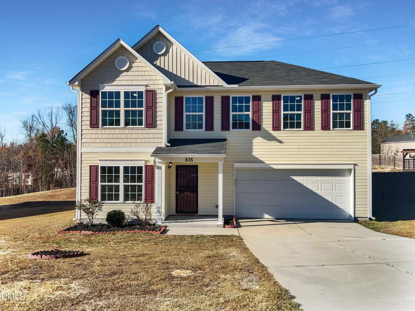 Two-story beige house with red shutters, a black front door, white trim, and a double garage. The driveway is concrete, and the yard is mostly grass with small shrubs and minimal landscaping. Trees are in the background.