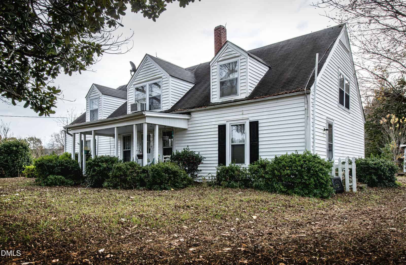 White two-story house with black shutters, front porch, large windows, dormers, and a brick chimney located at 120 Furney Pearce Road in Zebulon. The yard features bushes and trees, with a ground covering of dry leaves and grass.