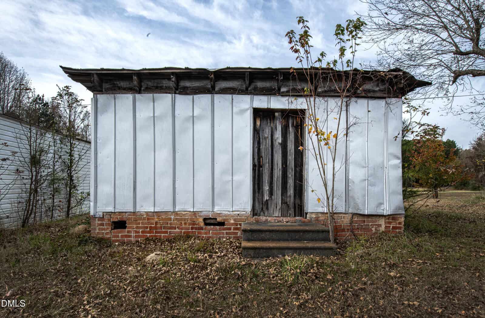 A small shed with metal siding, a brick foundation, and a weathered wooden door sits in a yard with fallen leaves. Steps lead to the entrance; a few young trees grow near the building. Sky is partly cloudy.