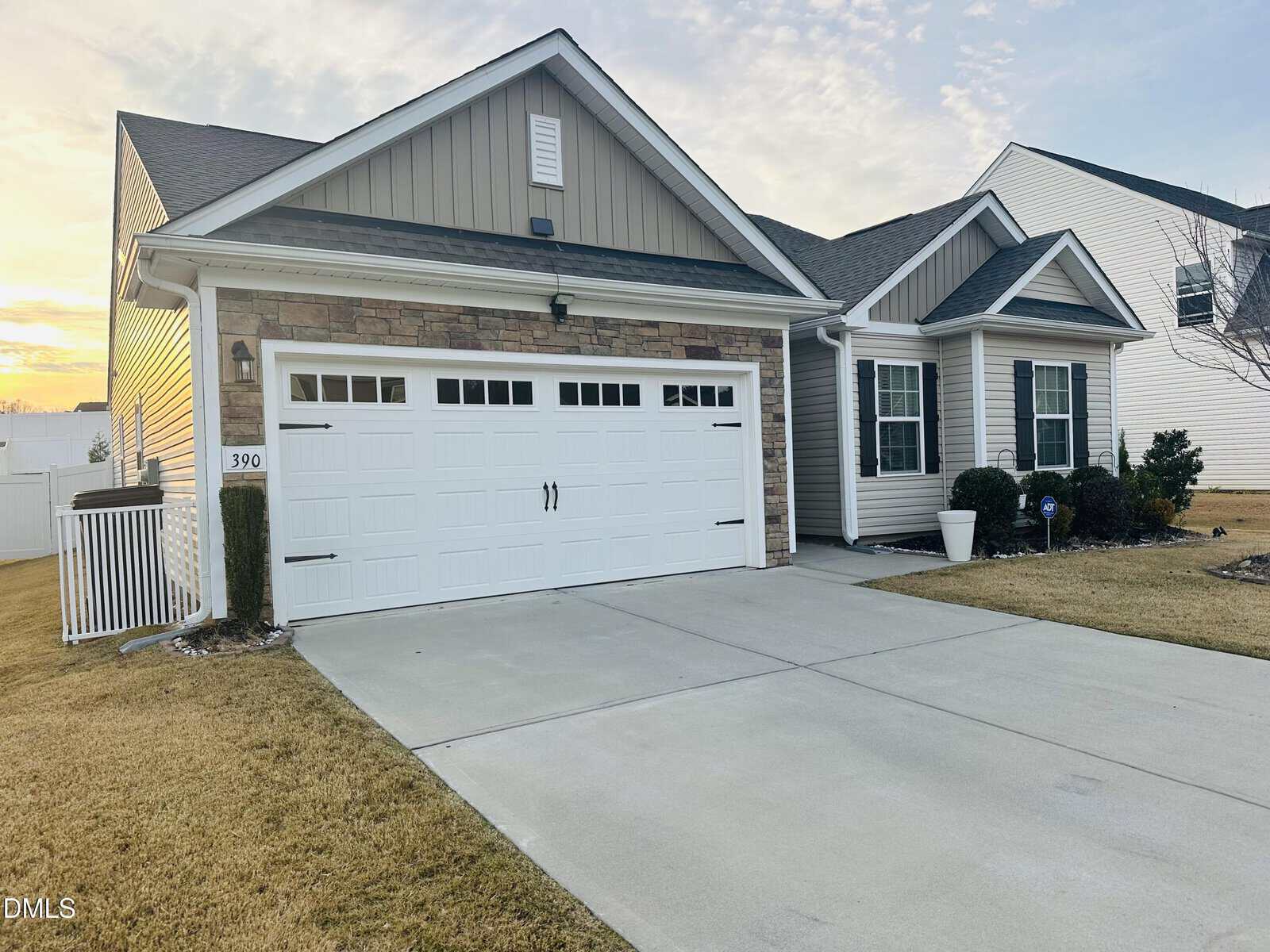 Single-story suburban house with stone and beige siding, a white double garage door, and manicured front yard. The driveway is concrete, and the sky is partly cloudy during sunset.