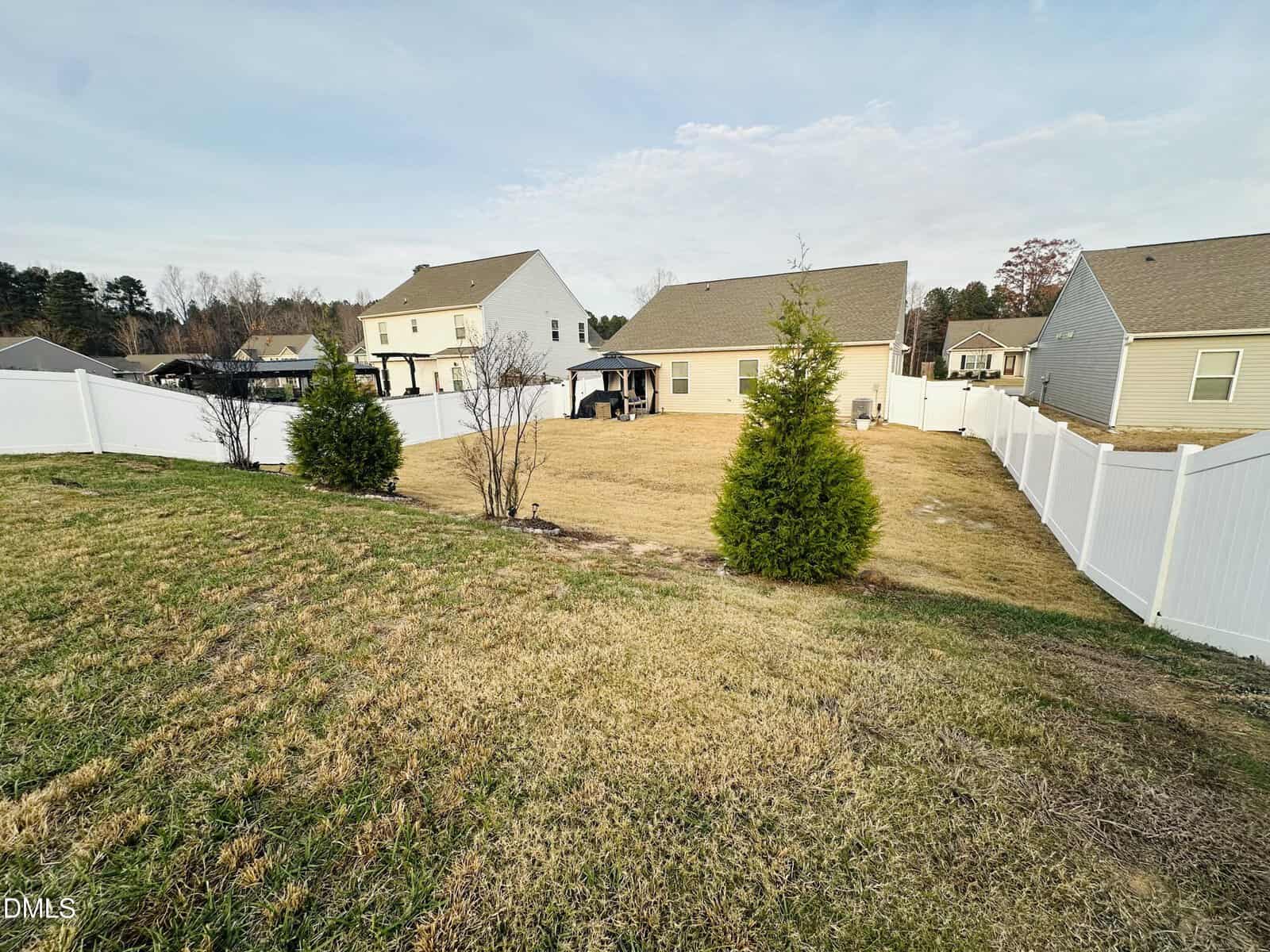 A fenced backyard with a grassy lawn, a few small trees, and neighboring houses in the background under a partly cloudy sky.