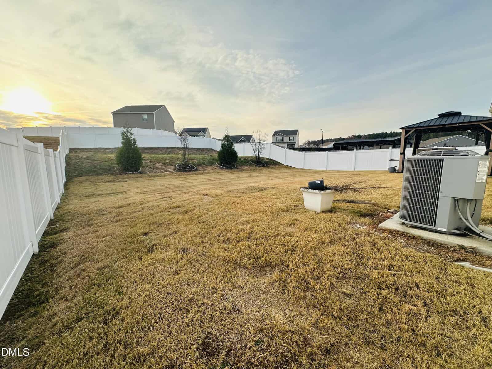 A fenced backyard with yellowish grass, a few small trees near the fence, and an outdoor air conditioning unit on a concrete slab. Houses and a setting sun are visible in the background.