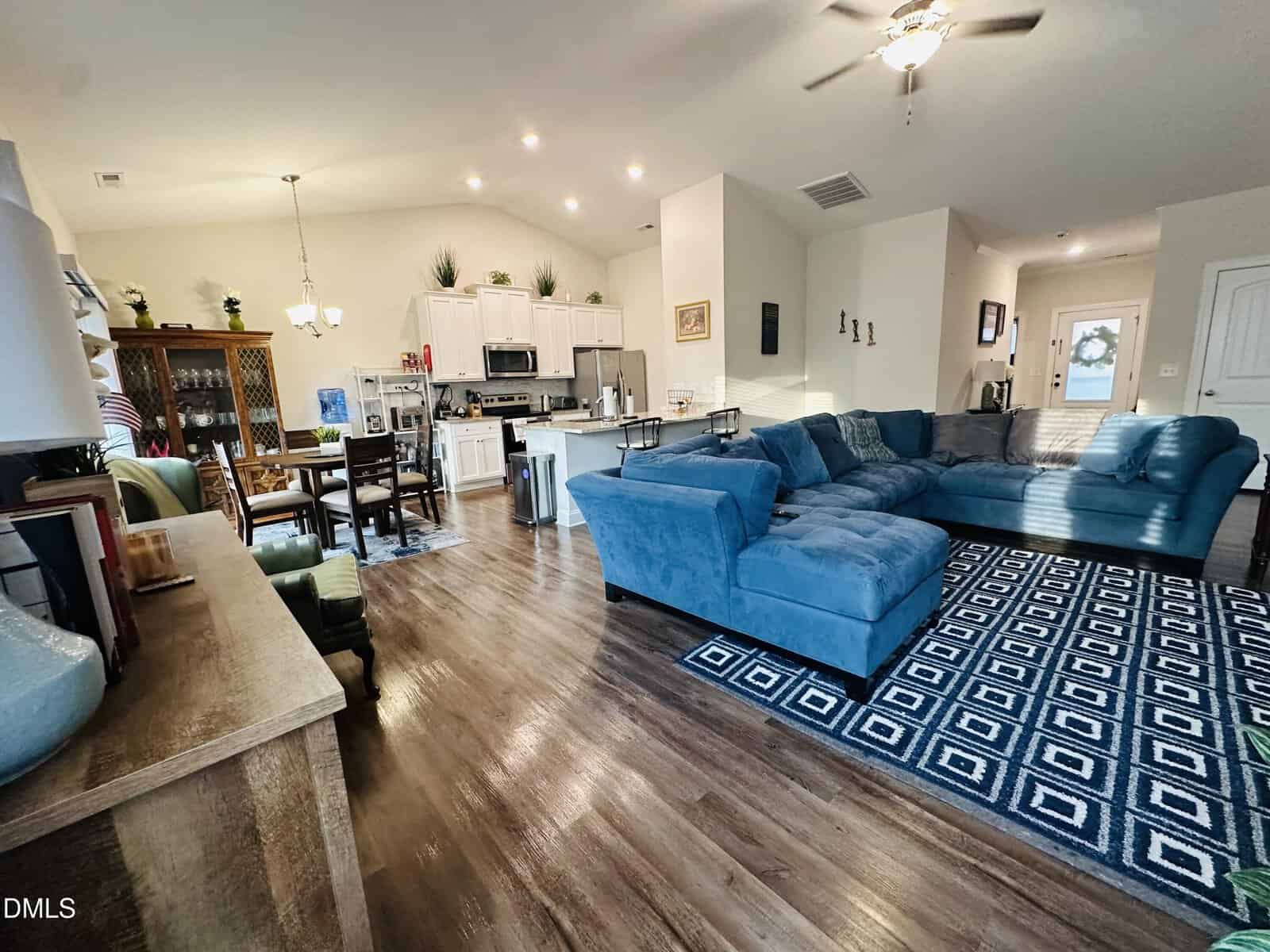 Open-concept living space with wood floors, a blue sectional sofa on a geometric rug, a dining area with a wooden table, a china cabinet, and a modern kitchen with white cabinets and stainless steel appliances.