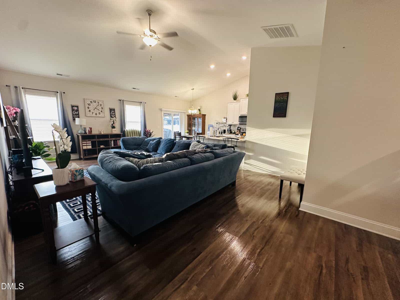 Open living room with a blue sectional sofa, flat-screen TV, houseplants, wood floors, and a view into a bright kitchen with white cabinets and dining table under vaulted ceiling with ceiling fans. Natural light fills the space.
