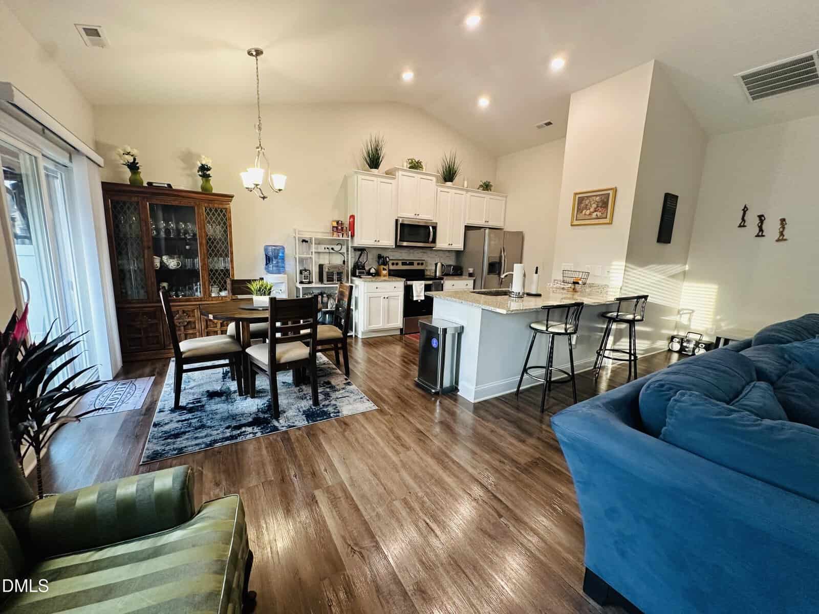 Open-concept kitchen and dining area with white cabinets, island with barstools, wooden floor, dining table, blue sofa, green striped chair, and decorative plants, all well-lit by natural and artificial light.