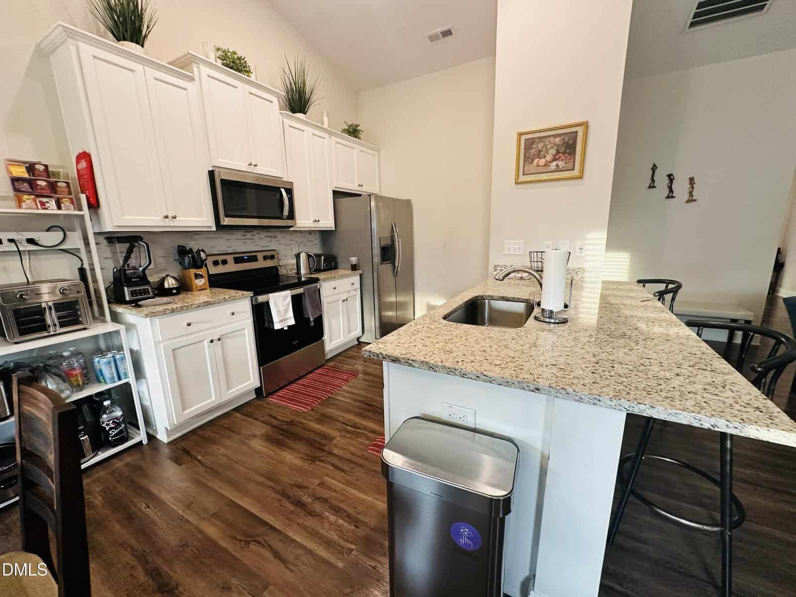 Modern kitchen with white cabinets, stainless steel appliances, granite countertops, and a breakfast bar with stools. Hardwood floors, wall art, and decorative plants add a cozy touch.