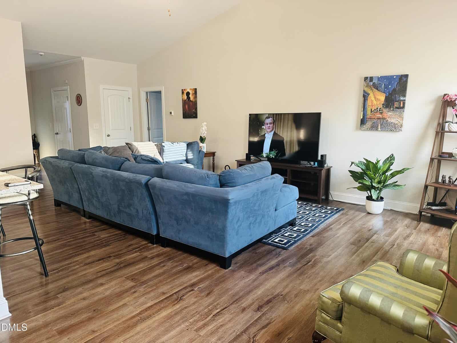 A spacious living room with a large blue sectional sofa, wooden floors, a TV on a stand, potted plants, wall art, a green armchair, and natural light streaming in from the left.