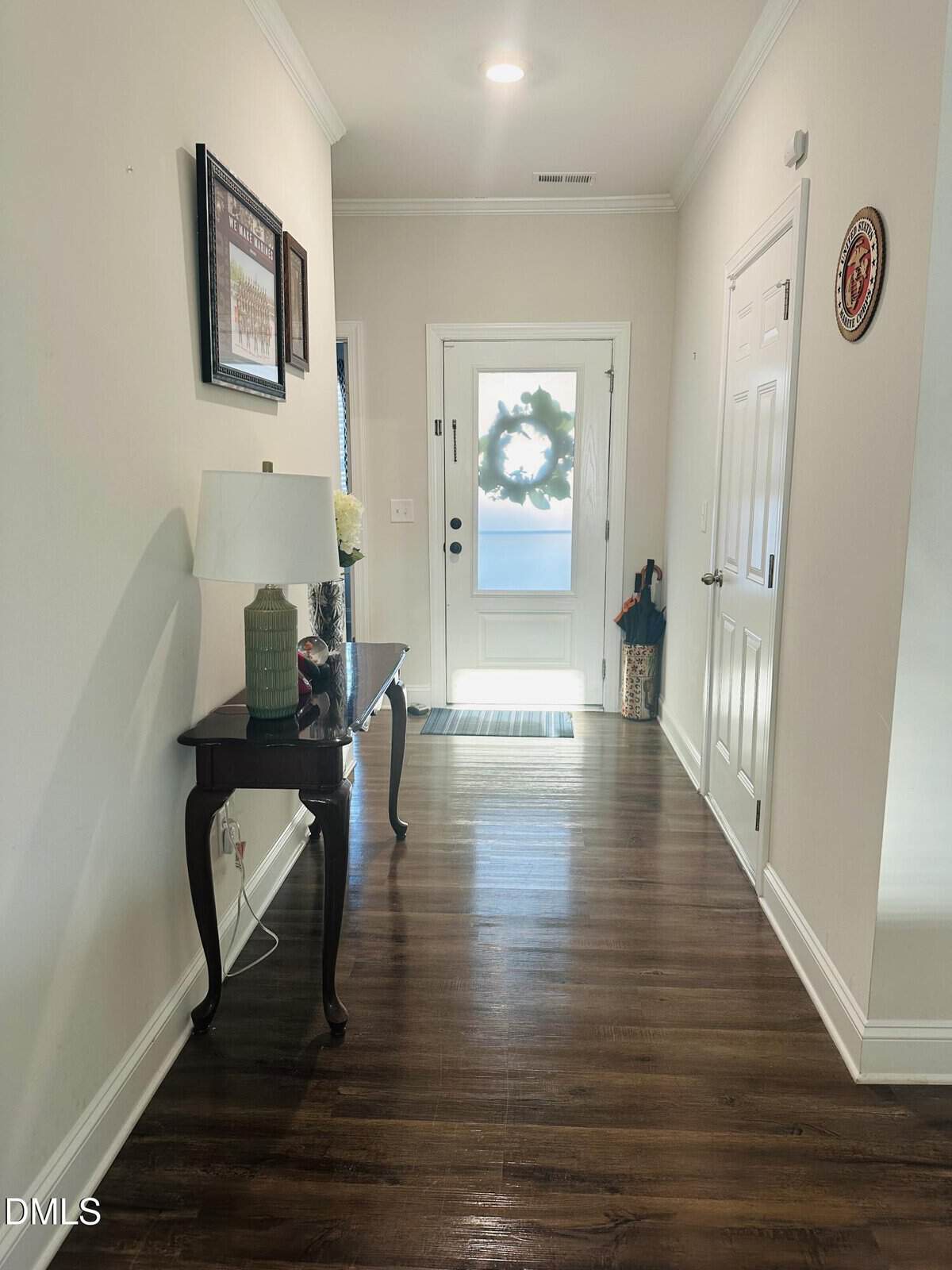 Bright hallway with wood floors, a small table holding a lamp and vase, framed art on the wall, and a white front door with a wreath. A striped rug lies in front of the door.