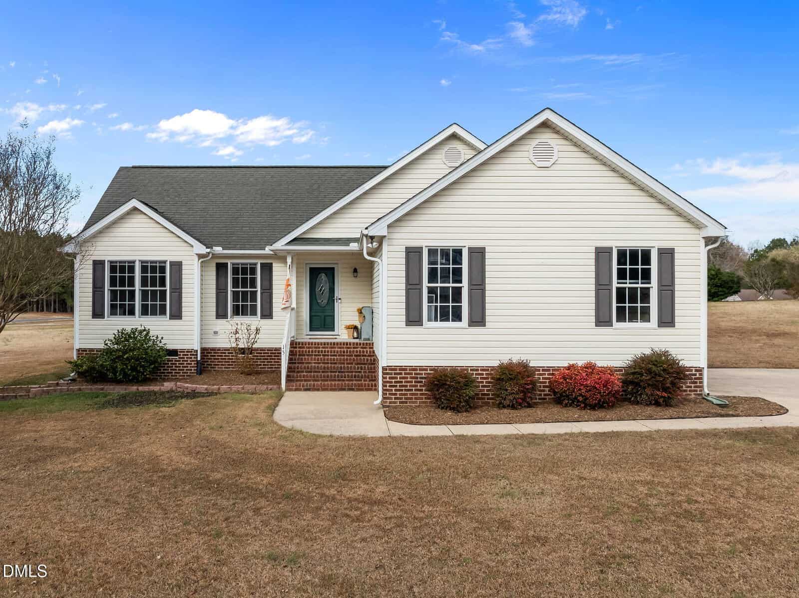Single-story white house with gray shutters, brick foundation, green shingle roof, front porch with chair, and simple landscaping, situated on a large, grassy lot under a partly cloudy sky.