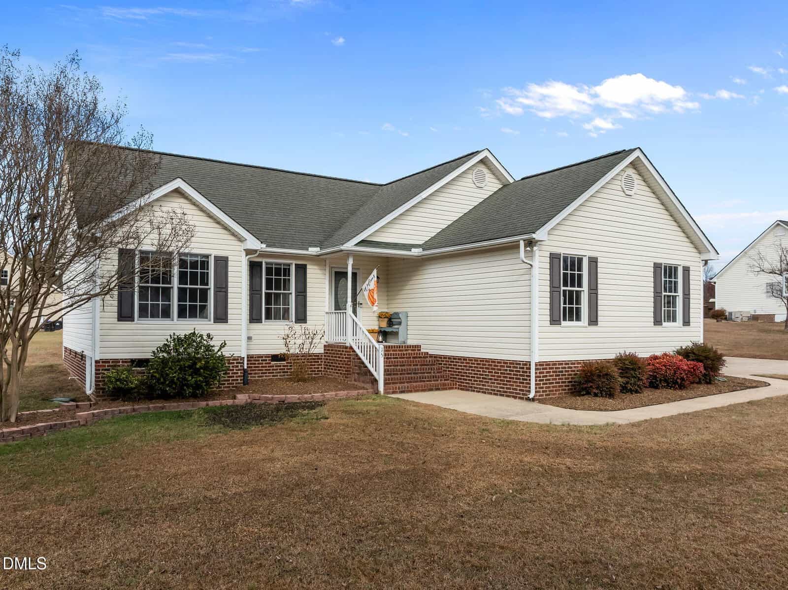 Charming single-story white house with gray roof and brick foundation located at 15 Woodcrest Drive, Youngsville. Features a small front porch with railing, flag, and steps. The yard is mostly brown grass with a few bushes and a tree, set under a clear sky with some clouds.
