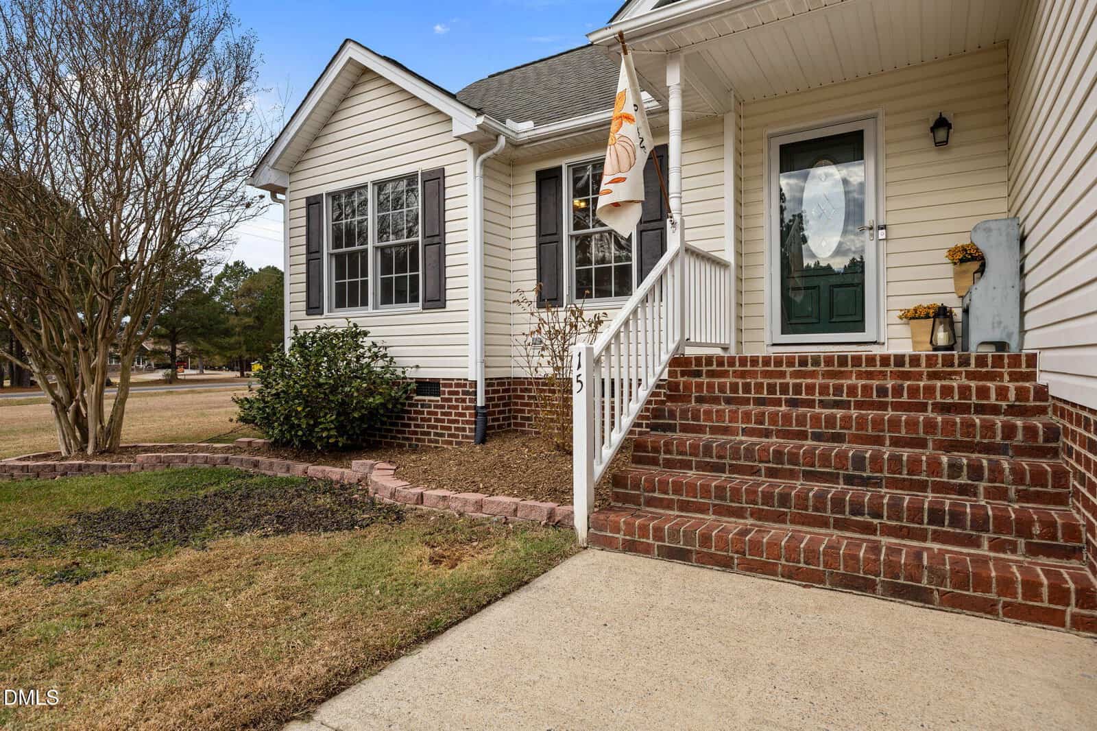 Front view of a house with white siding, dark shutters, a green door, brick steps, railing, potted plants, and a small yard with shrubs and a tree. House number 145 is displayed on the railing.