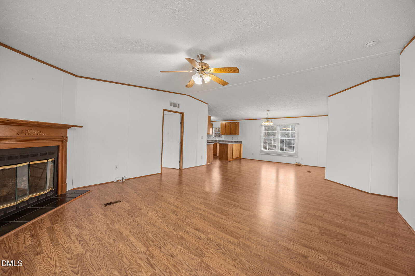 Spacious living area with wood-style flooring, a ceiling fan, a fireplace with a wooden mantel on the left, and an adjacent open kitchen and dining area visible in the background.