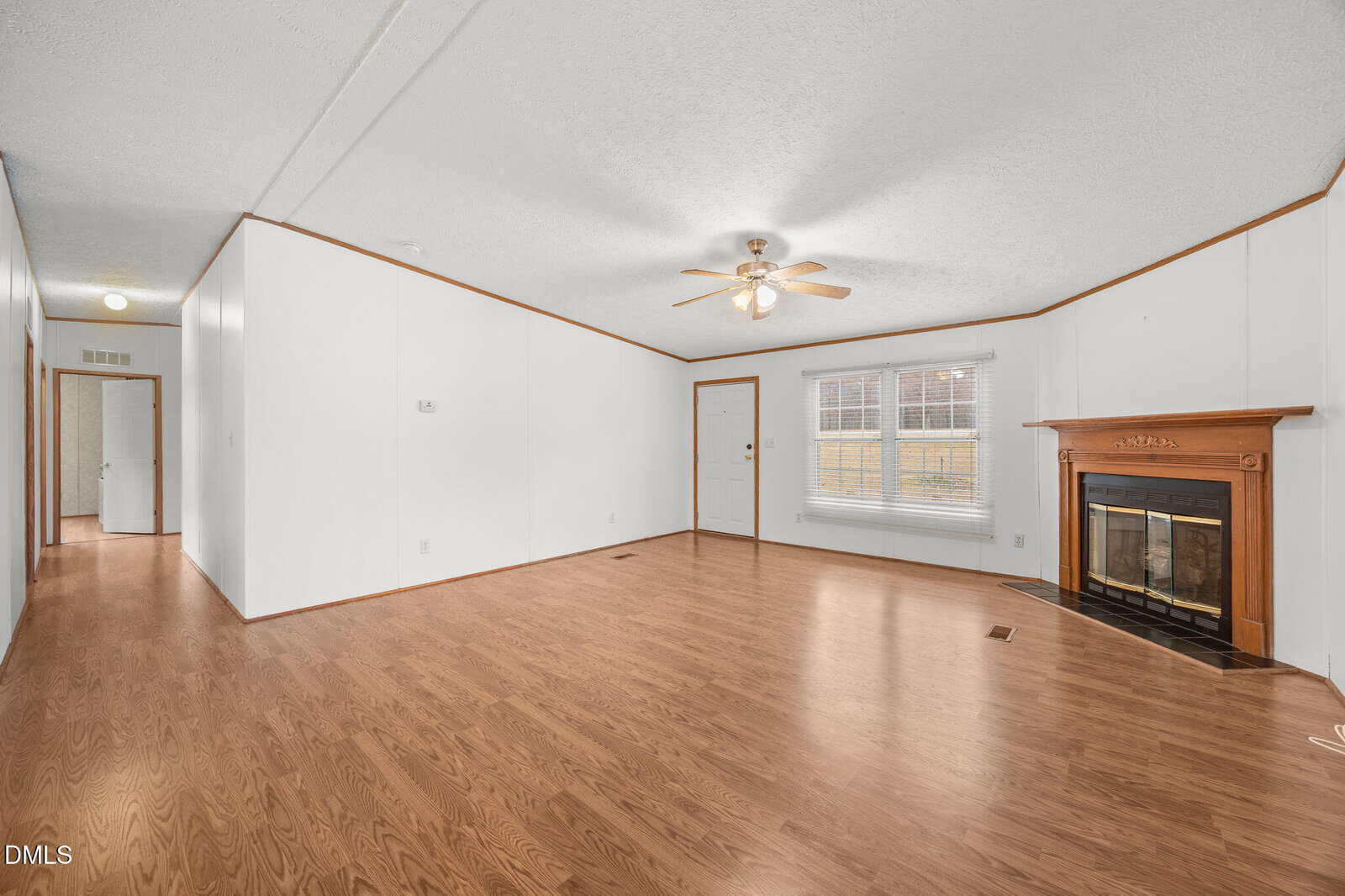 A spacious living room with white walls, wood-look flooring, a ceiling fan with light, a wooden fireplace, and a large window. A hallway and a front door are visible on the left side of the room.