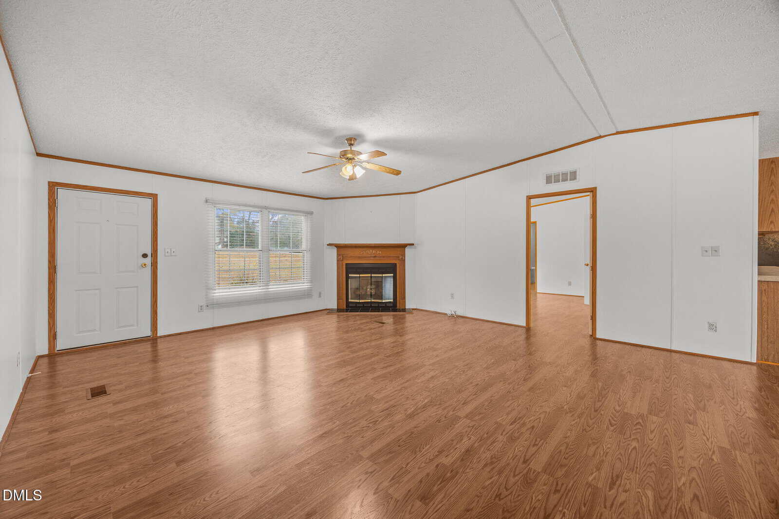 A spacious living room with light wood flooring, white walls, a ceiling fan with lights, a corner fireplace, large window, and open doorways leading to other areas of the house.