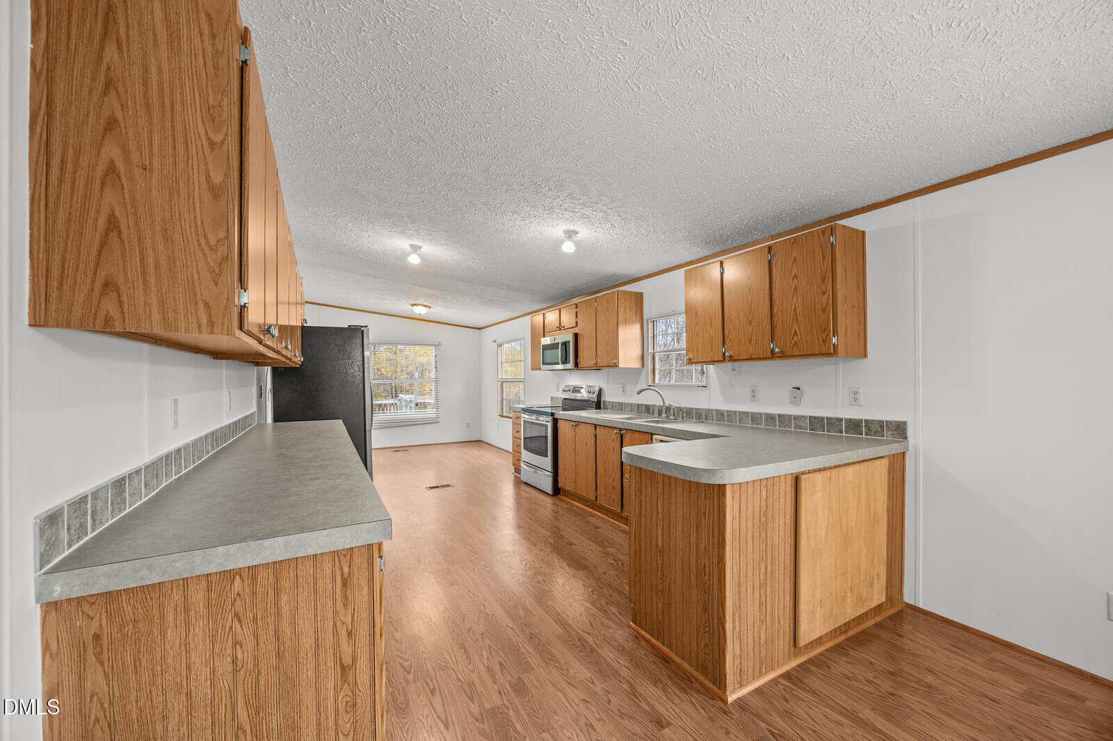 Kitchen with wood cabinets, laminate countertops, electric stove, dishwasher, and double sink. Wood-look flooring throughout, with two windows providing natural light at the far end of the room.