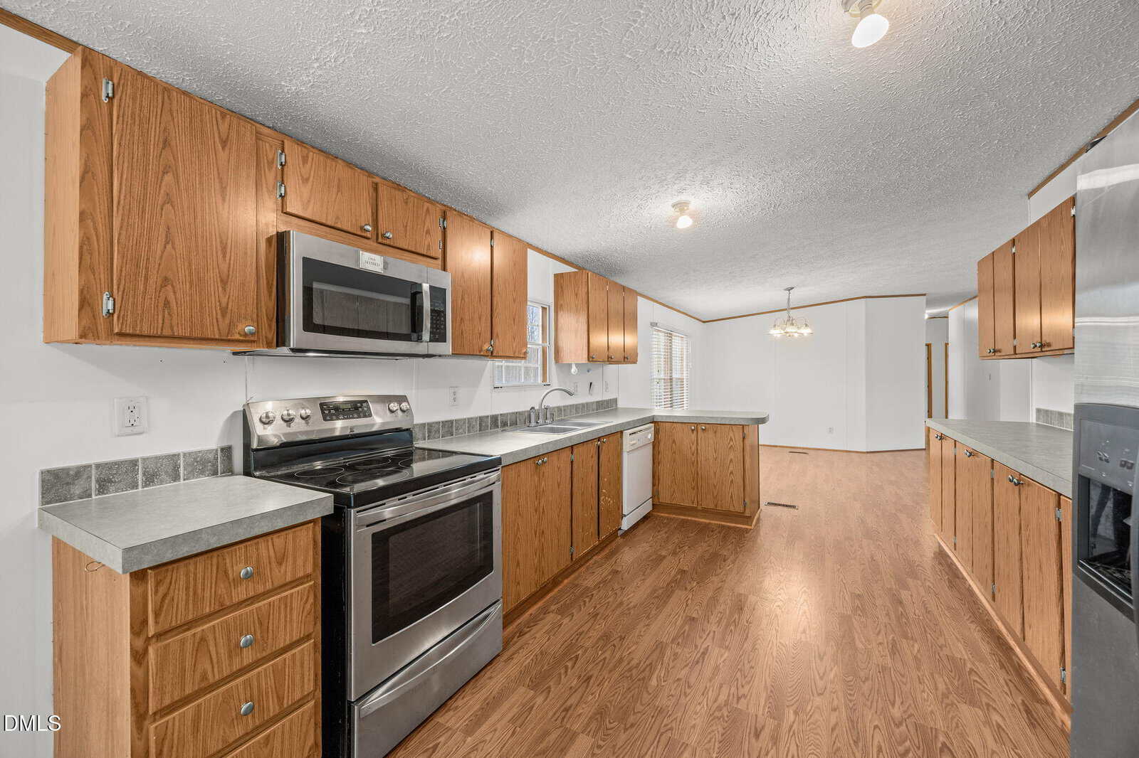 A spacious kitchen with wood cabinets, stainless steel appliances, gray countertops, and hardwood flooring. The kitchen opens into a large, well-lit dining and living area with neutral-colored walls.