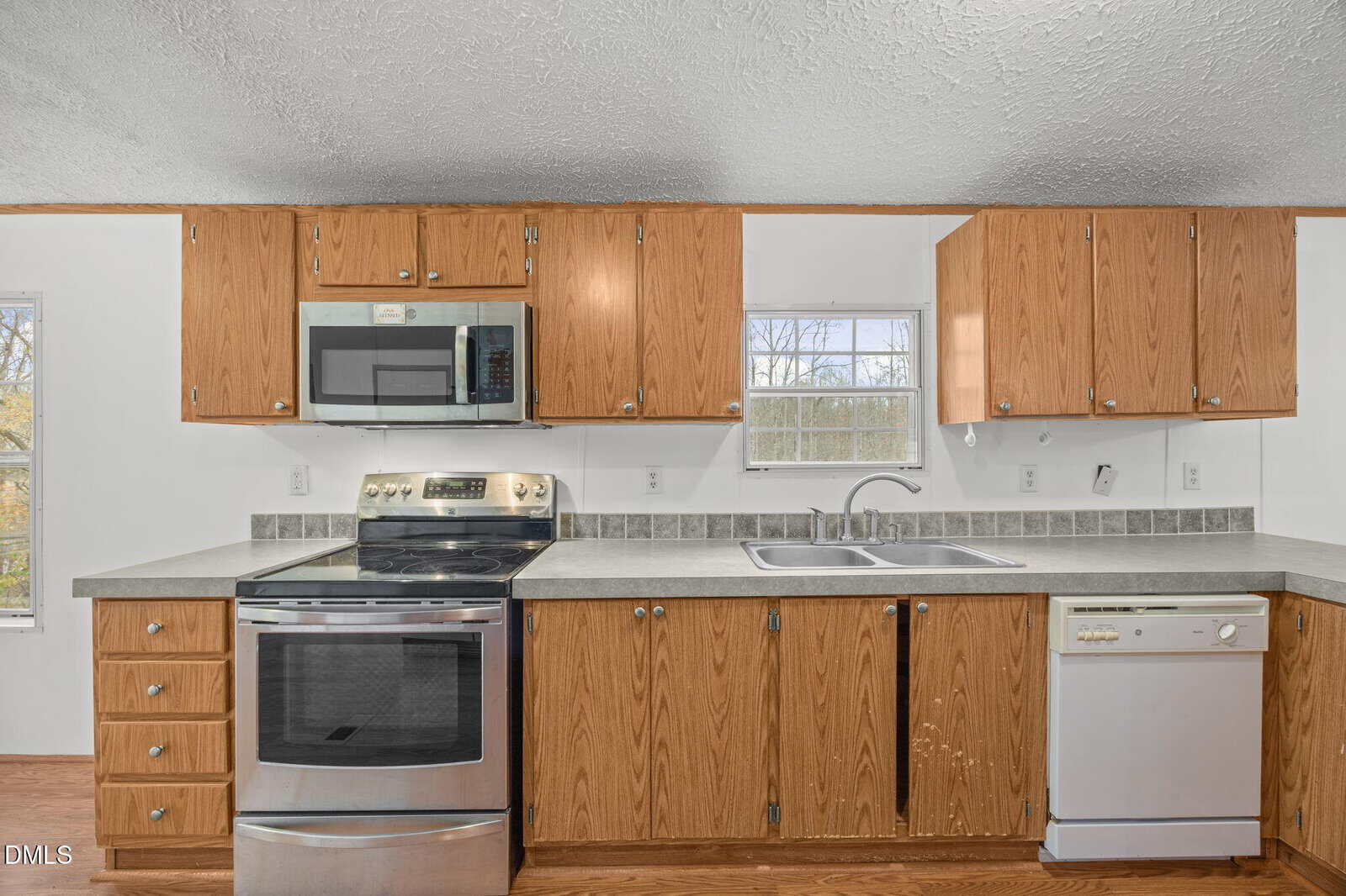 A kitchen with wood cabinets, a stainless steel electric stove and oven, a built-in microwave, a double sink under two windows, and a white dishwasher, with neutral countertops and tile backsplash.