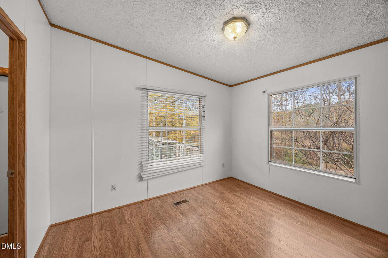 A small, empty room with white walls, wood trim, and wood laminate flooring. Two large windows let in natural light. A ceiling light fixture is visible at the center of the ceiling.