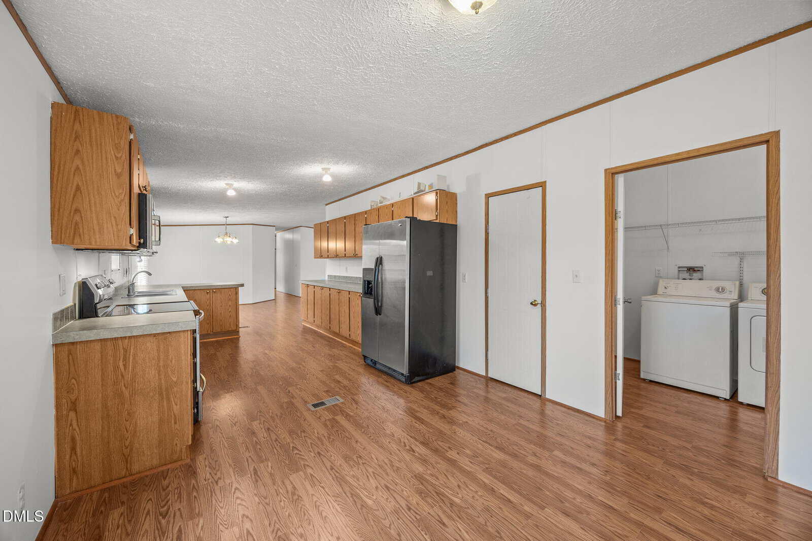 A spacious kitchen with wood cabinets, stainless steel appliances, and laminate wood flooring. A laundry room with a washer and dryer is visible through an open door on the right. The walls are white with wood trim.