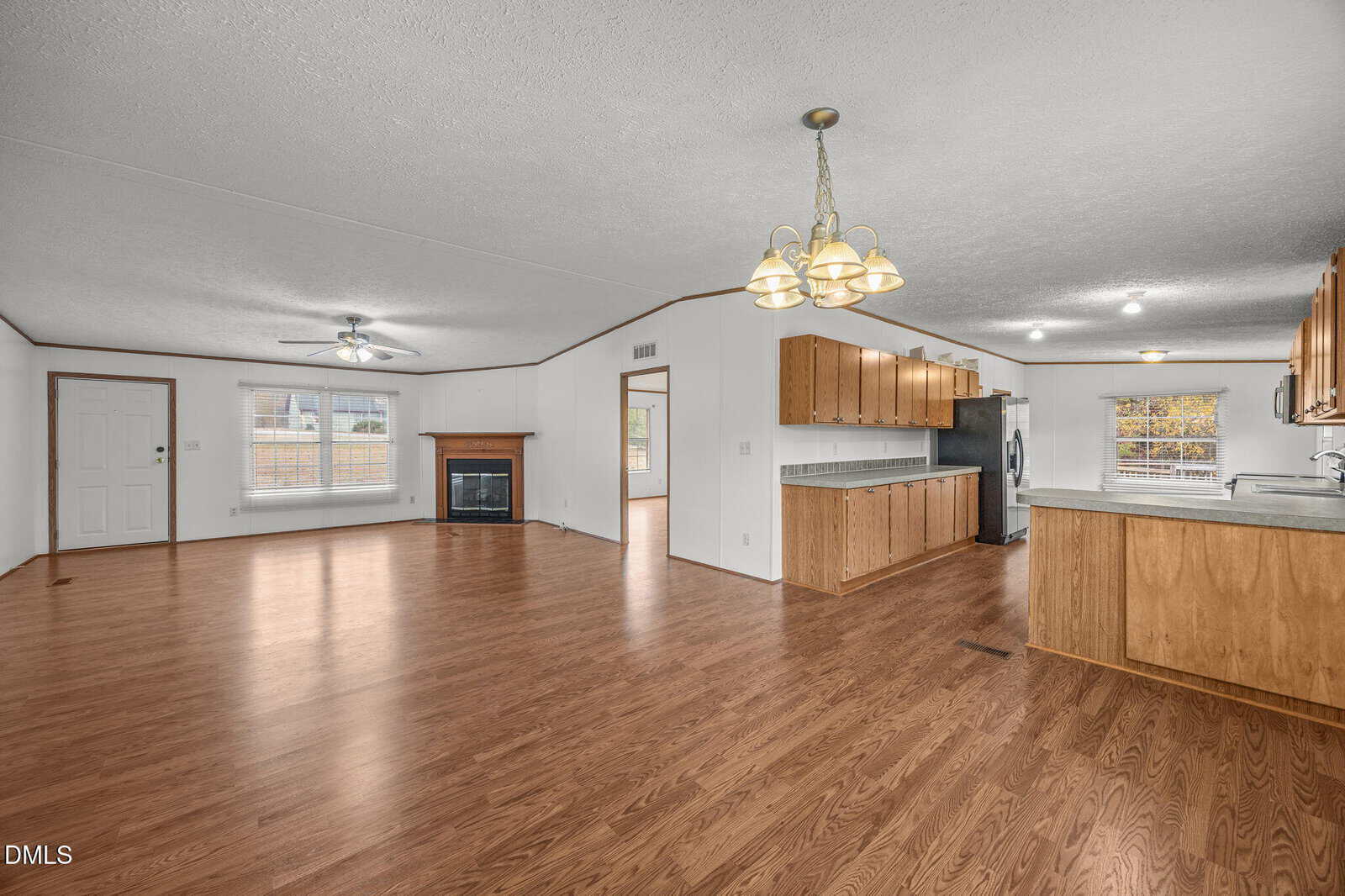 Open-concept room with wood flooring, a kitchen with wooden cabinets and stainless steel appliances, a dining area with a chandelier, and a living area featuring a fireplace and large windows.
