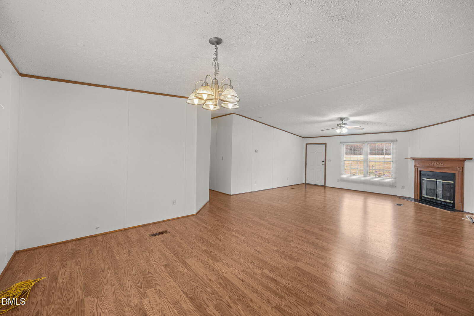 Spacious empty living room with wood flooring, white walls, a ceiling fan, chandelier, and a fireplace by a large window; broom on the floor in the corner.