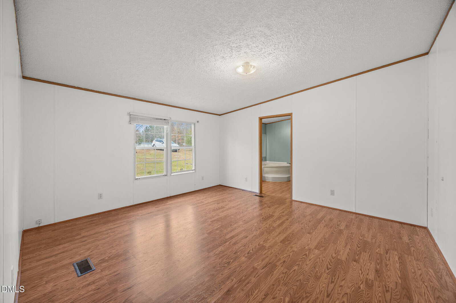 Empty room with wood flooring, white walls, a window with a view of grass, and a ceiling light fixture. An open doorway leads to a bathroom with a tub visible.