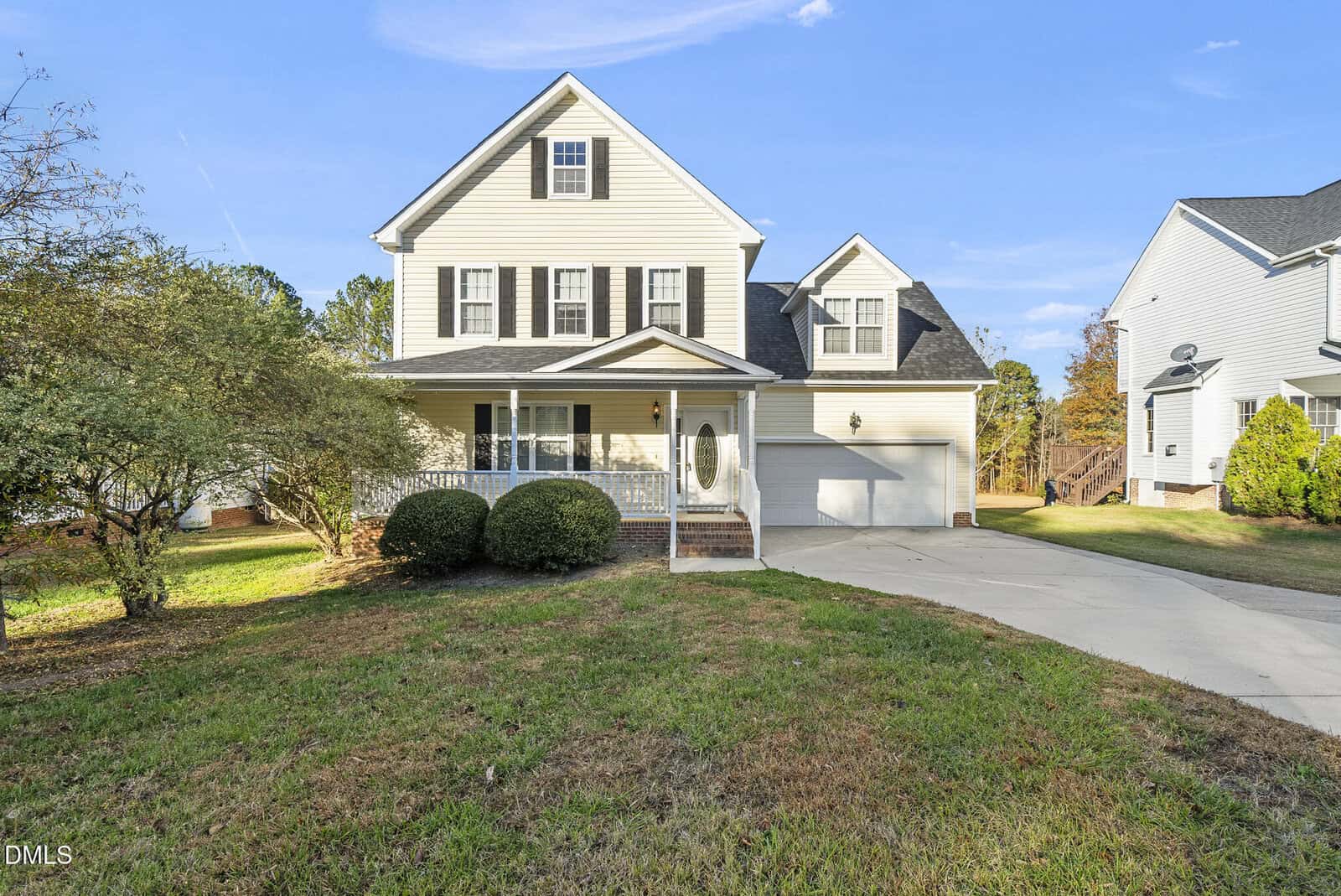 This two-story beige house at 1589 Rogers Pointe Lane in Creedmoor features black shutters, a covered front porch, an attached two-car garage, a grassy lawn, a driveway, and a small tree on the left side.
