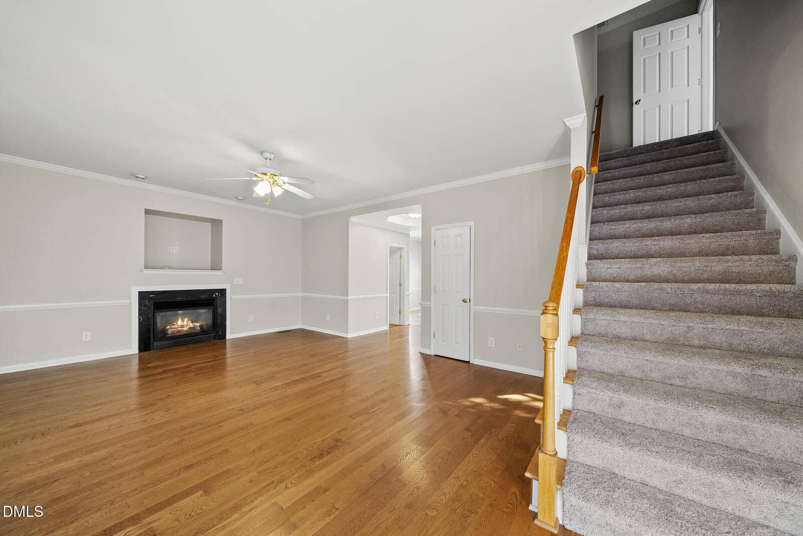 A spacious living room with light beige walls, wood flooring, a ceiling fan, a corner fireplace, and a staircase with carpeted steps and a wooden handrail leading to an upper floor.