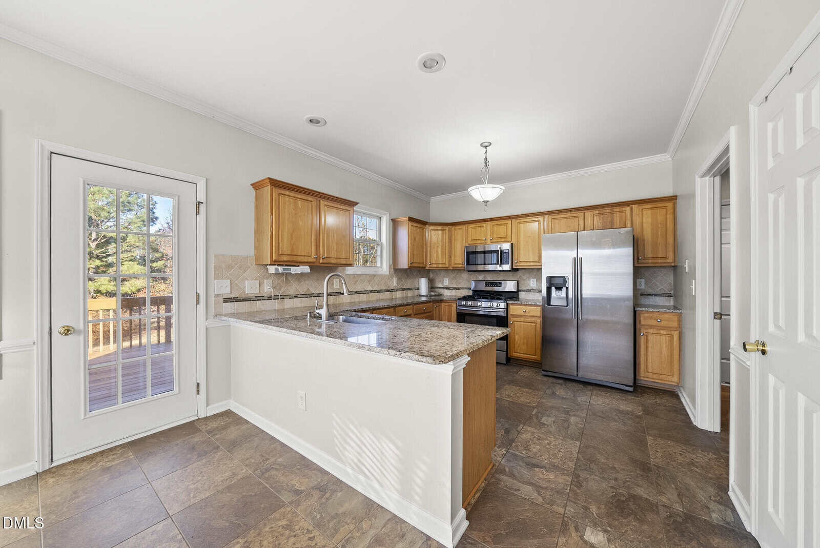 A spacious kitchen with wooden cabinets, stainless steel appliances, marble countertops, a double-door refrigerator, a central island with a sink, tile flooring, and a door leading to an outdoor deck.