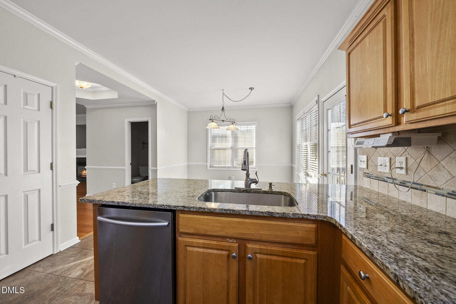 Bright kitchen with granite countertops, wood cabinets, a built-in sink, black dishwasher, tile floor, and large windows letting in natural light. Dining area and white doors visible in the background.