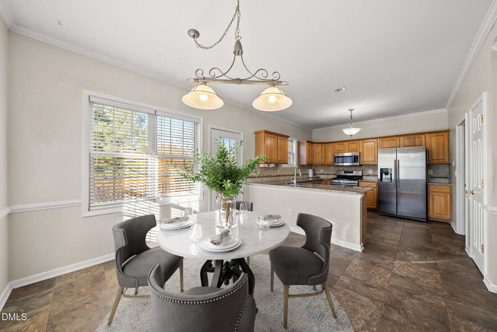 Bright kitchen with wood cabinets, stainless steel appliances, a marble dining table set for four, gray chairs, a window with blinds, and a decorative hanging light fixture above the table.