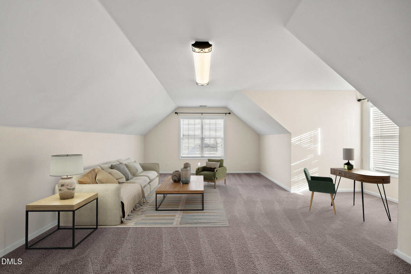 A bright, spacious attic room with sloped ceilings, a beige sectional sofa, coffee table, two green chairs, a desk, and windows letting in natural light. Neutral carpet covers the floor.