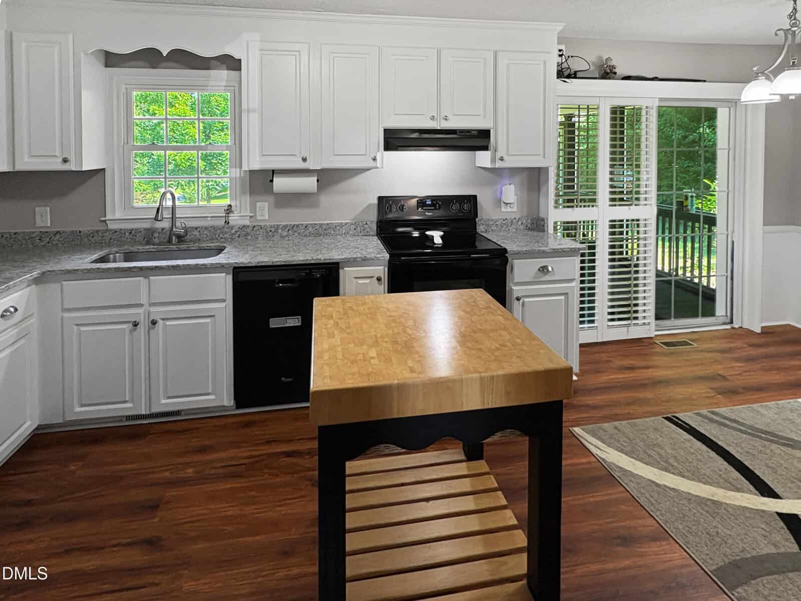 Bright kitchen with white cabinets, stainless sink, black appliances, a central wood-topped island, large window, sliding glass doors with blinds, and hardwood floors. A partial view of a rug is visible.