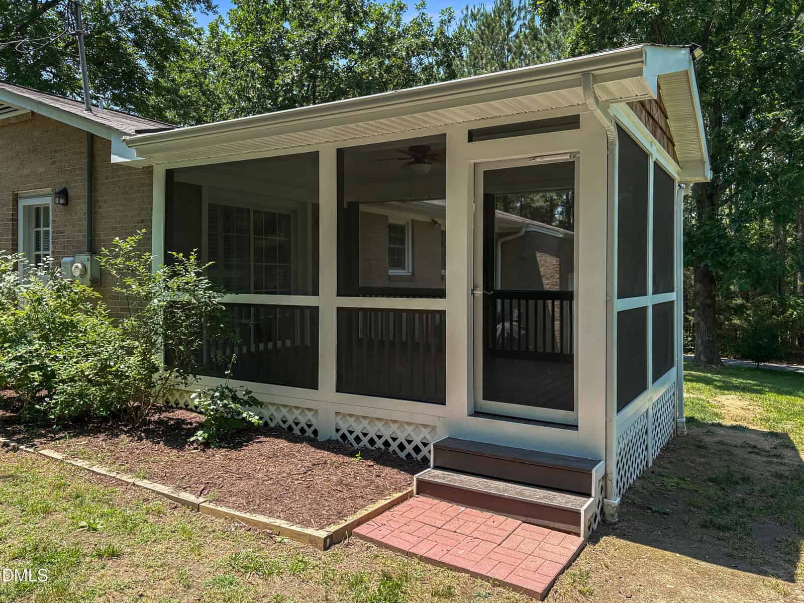 A screened-in porch with white framing and steps leads to the backyard of a brick house, surrounded by greenery and shaded by trees.