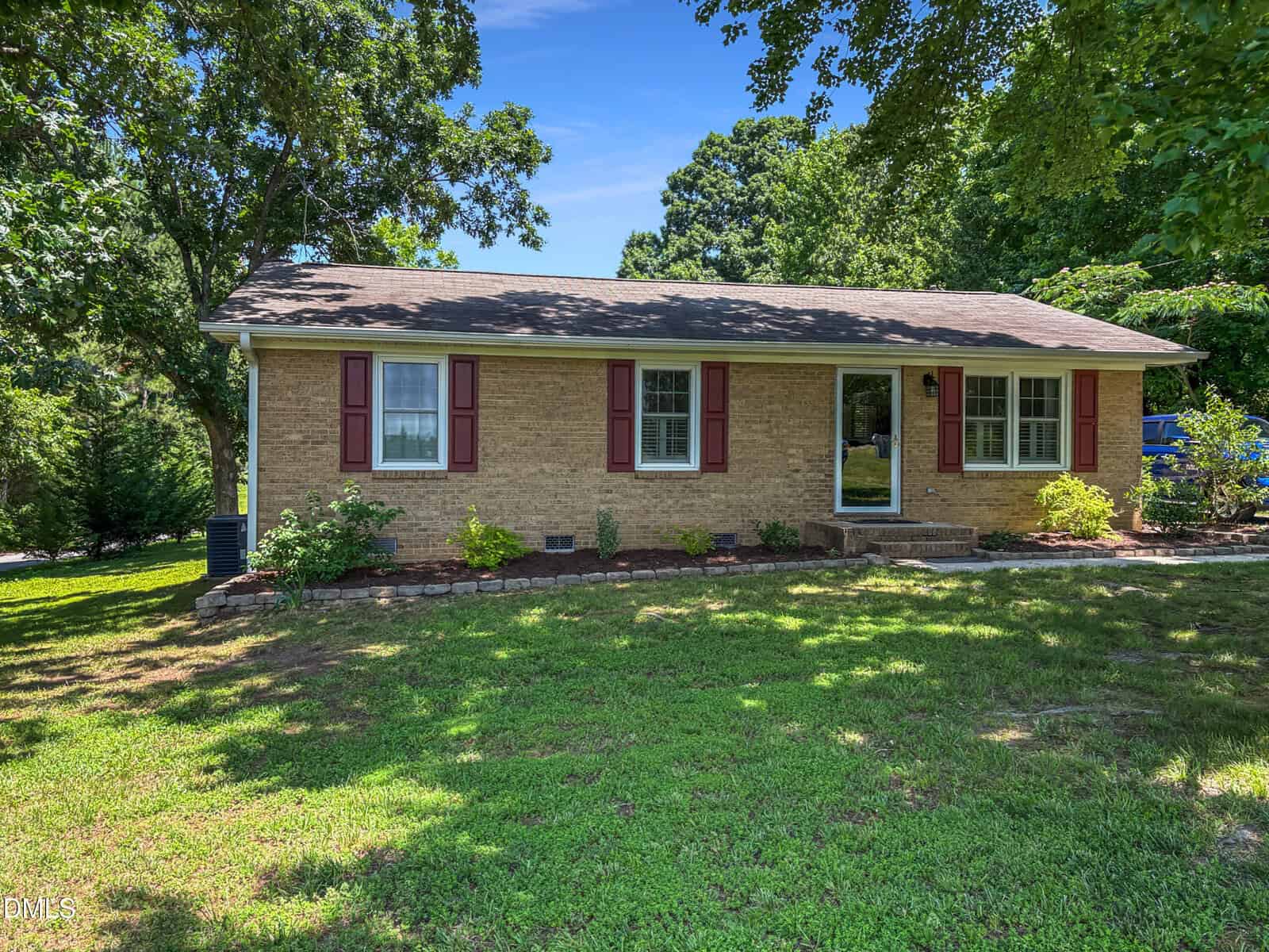 A single-story brick house at 573 Arnold Road in Louisburg features red shutters, white-framed windows, and a neatly landscaped front yard with green grass and trees under a clear blue sky.