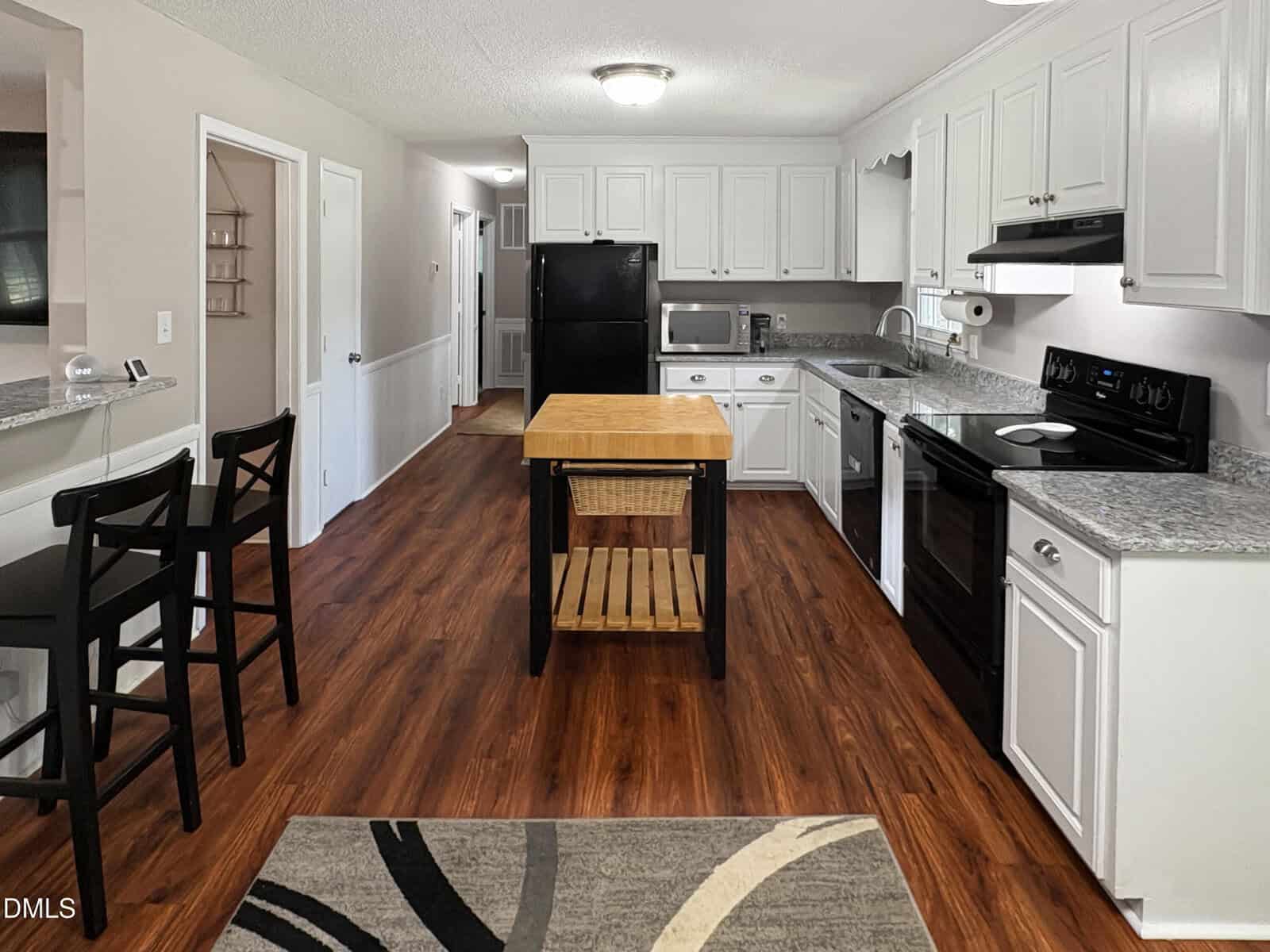 Modern kitchen with white cabinets, black appliances, wood floors, a central island with a wooden shelf, and a small dining area with two black chairs near a breakfast bar. A hallway is visible in the background.
