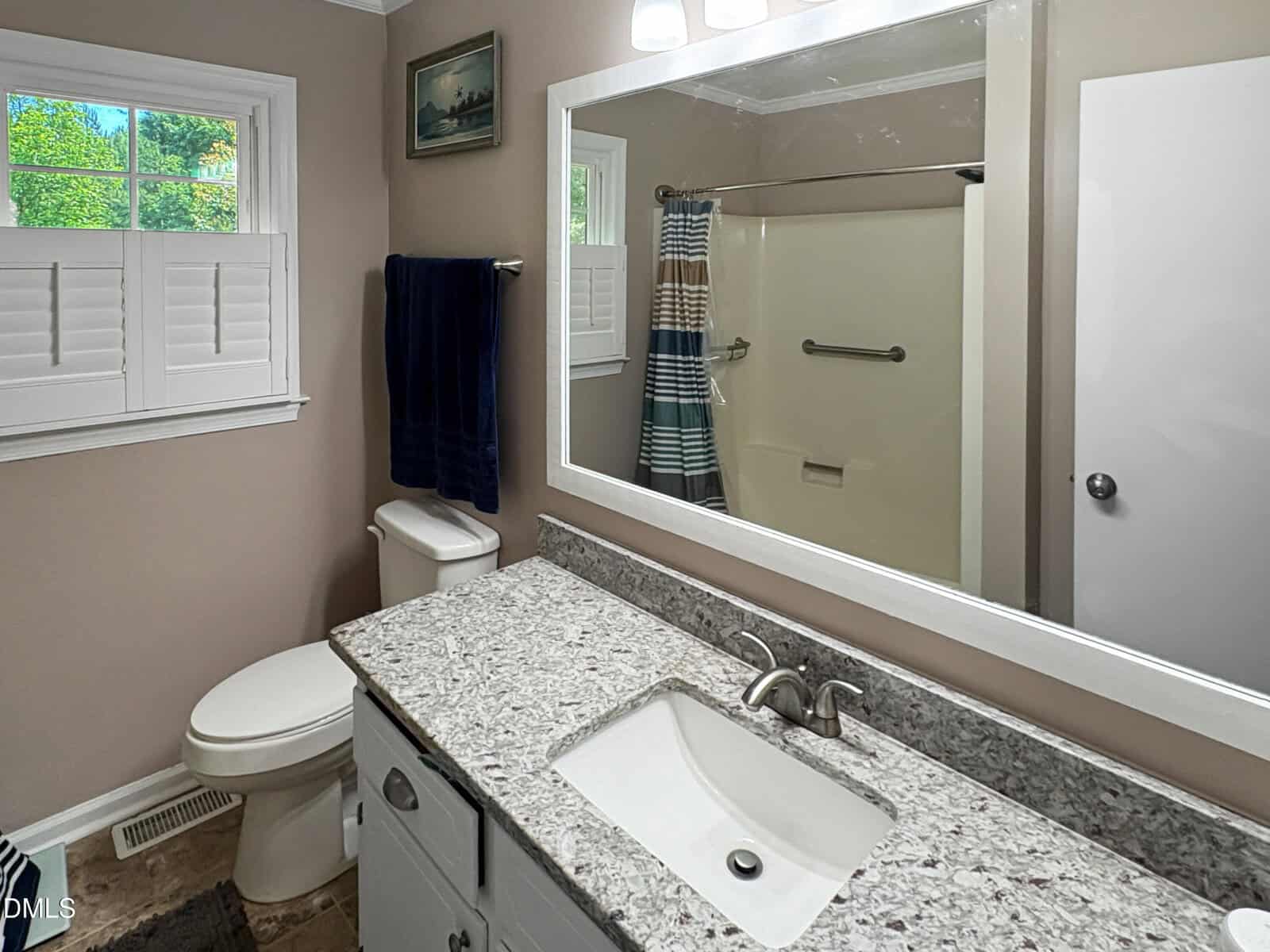 A bathroom with a granite countertop, white sink, toilet, towel rack, and a bathtub with a striped shower curtain; there’s a window with shutters and a mirror above the sink.